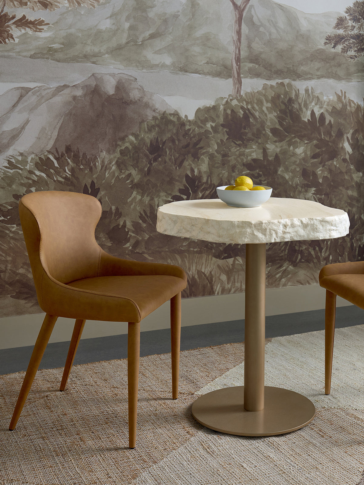 Wavy-edged cream bistro table between tan chairs, with a bowl of lemons, a woven rug and a landscape mural behind.