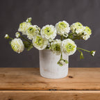 Several cream artificial flower sprays arranged in a pale cylindrical vase on a wooden table against a dark grey wall.