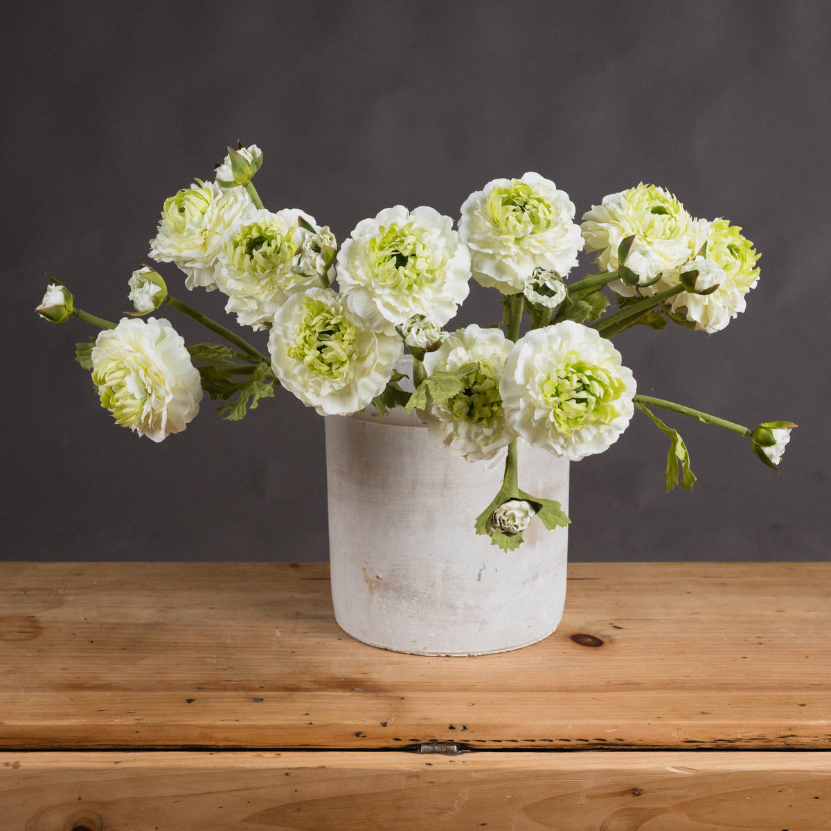 Several cream artificial flower sprays arranged in a pale cylindrical vase on a wooden table against a dark grey wall.