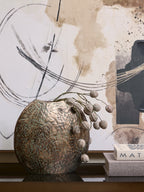 Weathered bronze-toned vase with pod-covered decorative stems trailing to the right, beside stacked books beneath abstract artwork.