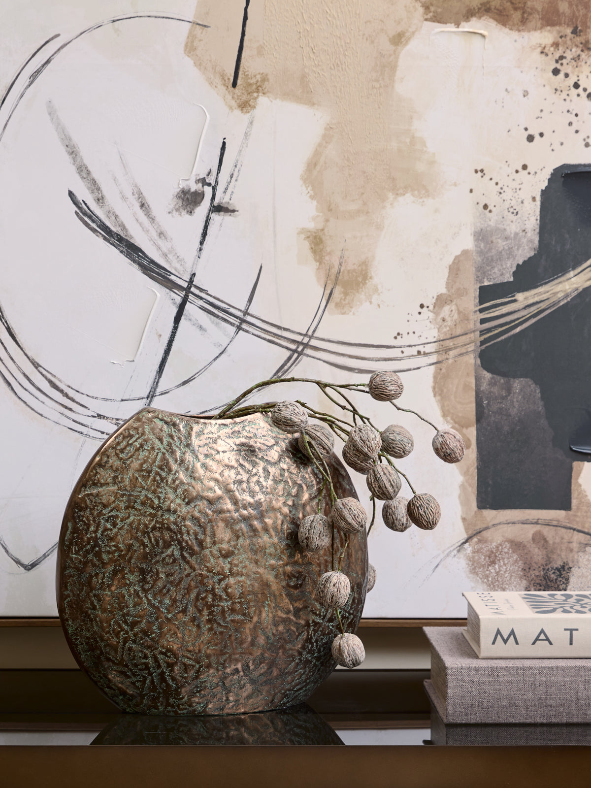 Weathered bronze-toned vase with pod-covered decorative stems trailing to the right, beside stacked books beneath abstract artwork.