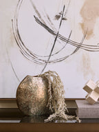 Weathered vase holding pale trailing stems on a reflective console beside stacked books and a geometric ornament, with abstract artwork behind.