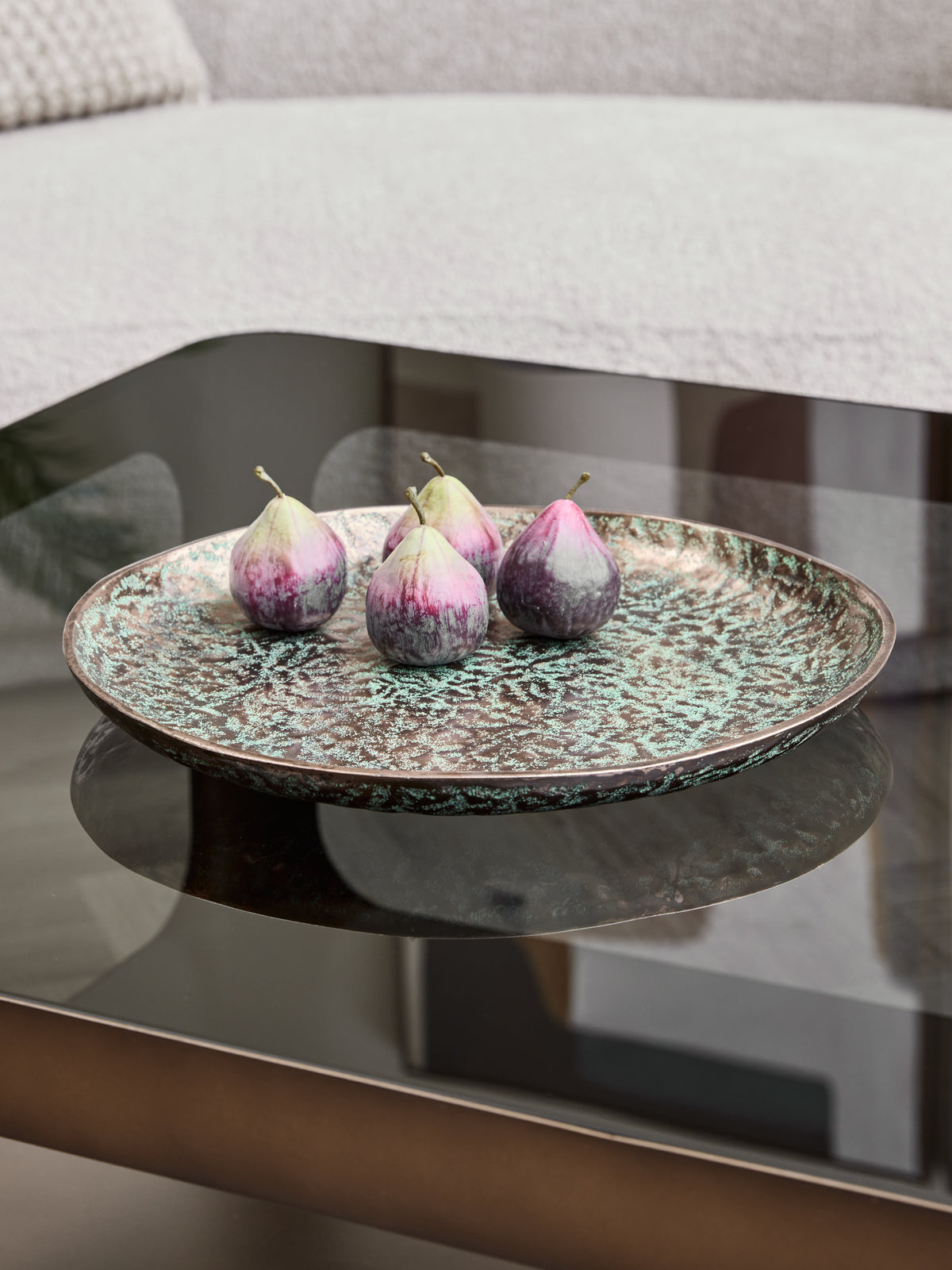 Bronze and verdigris platter holding four pear-shaped ornaments on a reflective coffee table beside a pale sofa.