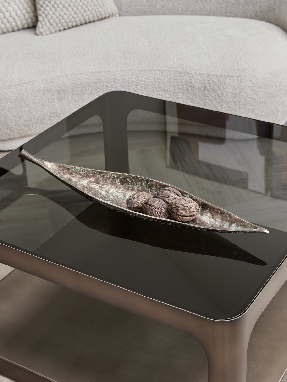 Leaf bowl holding four textured decorative spheres on a dark glass coffee table in front of a cream sofa.