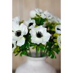 Close view of white anemone flowers with dark centres and green foliage above the rim of a white vase, with a softly blurred background.