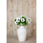 White anemone flowers arranged in a rounded white vase on a pale wooden surface against vertical wood panelling.