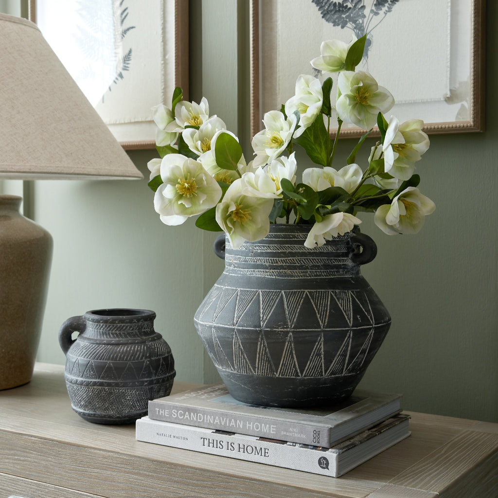 White artificial flowers arranged in a large dark patterned vase on stacked books, beside a smaller matching vessel and a lamp beneath framed botanical prints.