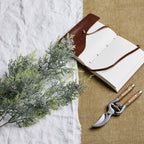 Frosted artificial conifer foliage laid diagonally across white cloth beside an open notebook with a brown cover and pruning shears on tan fabric.