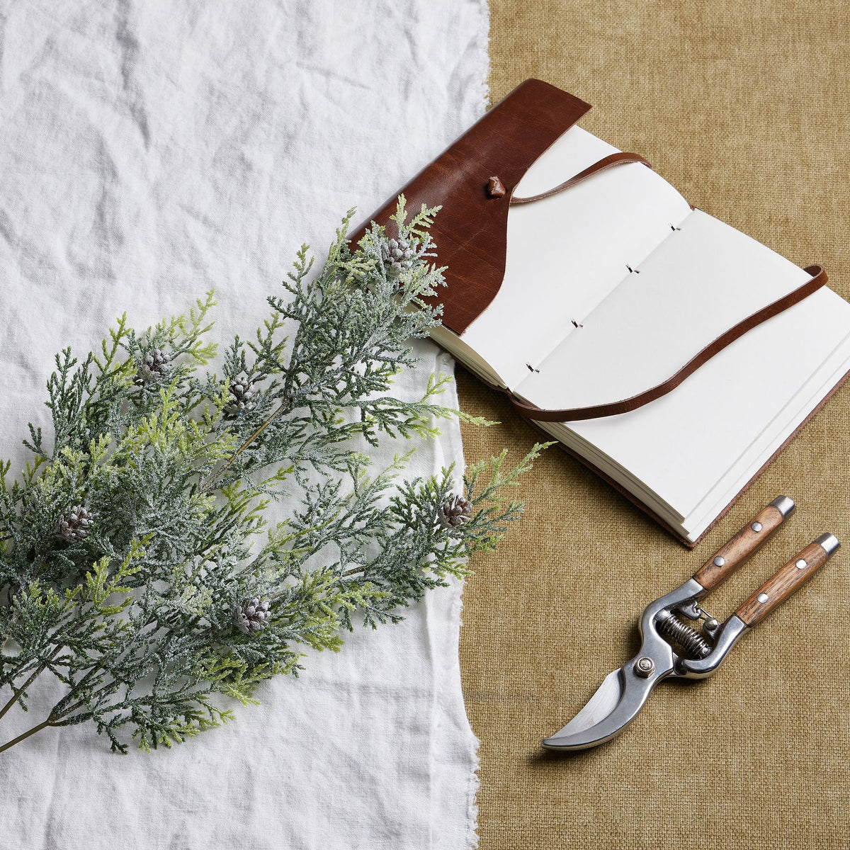 Frosted artificial conifer foliage laid diagonally across white cloth beside an open notebook with a brown cover and pruning shears on tan fabric.