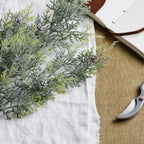 Close-up of white-frosted green conifer sprays and brown pinecones on white cloth, with part of an open notebook and pruning shears beside them.