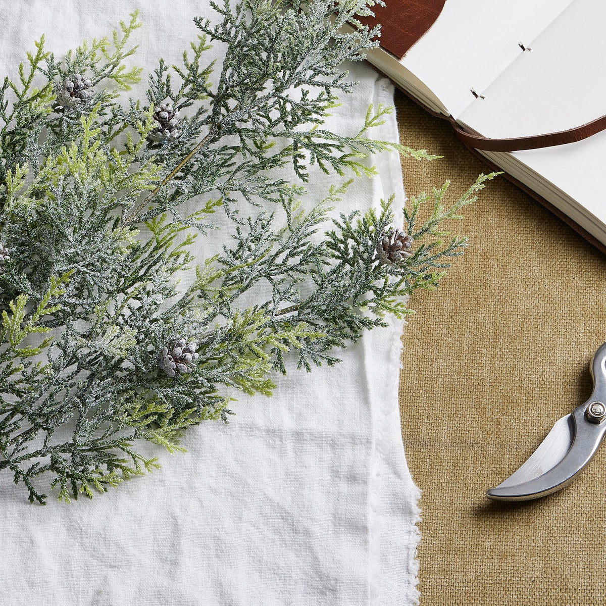 Close-up of white-frosted green conifer sprays and brown pinecones on white cloth, with part of an open notebook and pruning shears beside them.