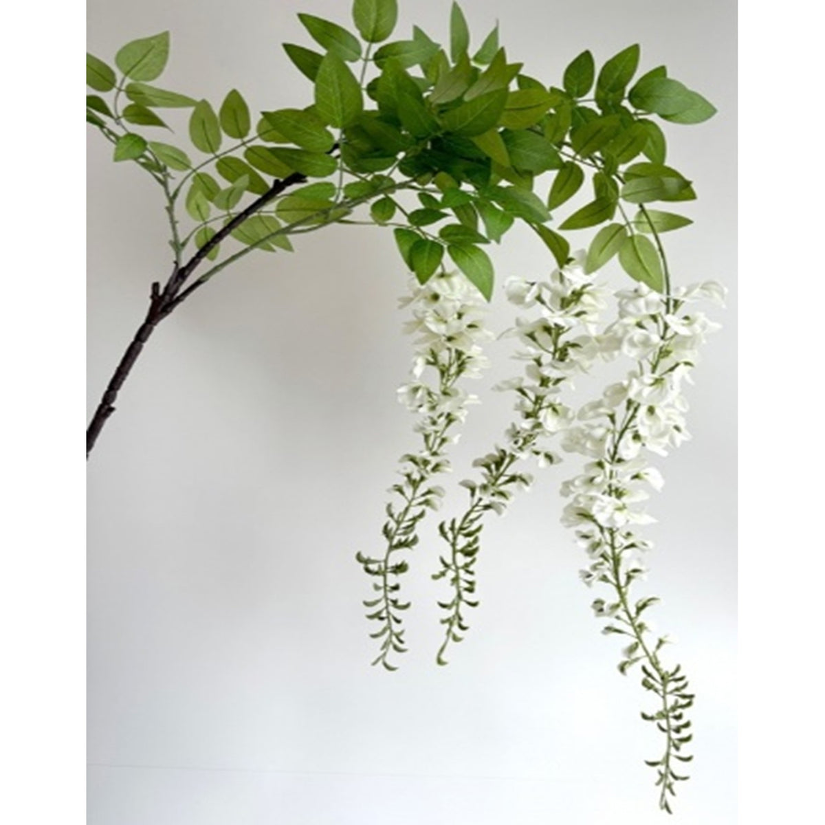 Close view of an artificial brown branch with green leaves above hanging clusters of white flowers and slender green tips against a pale background.