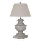 White-grey urn-shaped table lamp on a square plinth, with a pale tapered shade and ball finial against a white background.