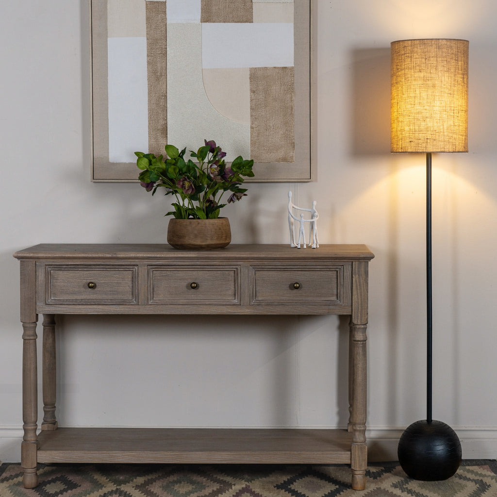 Floor lamp shown illuminated beside a wooden console, with neutral abstract artwork, flowers and a white figurine group.