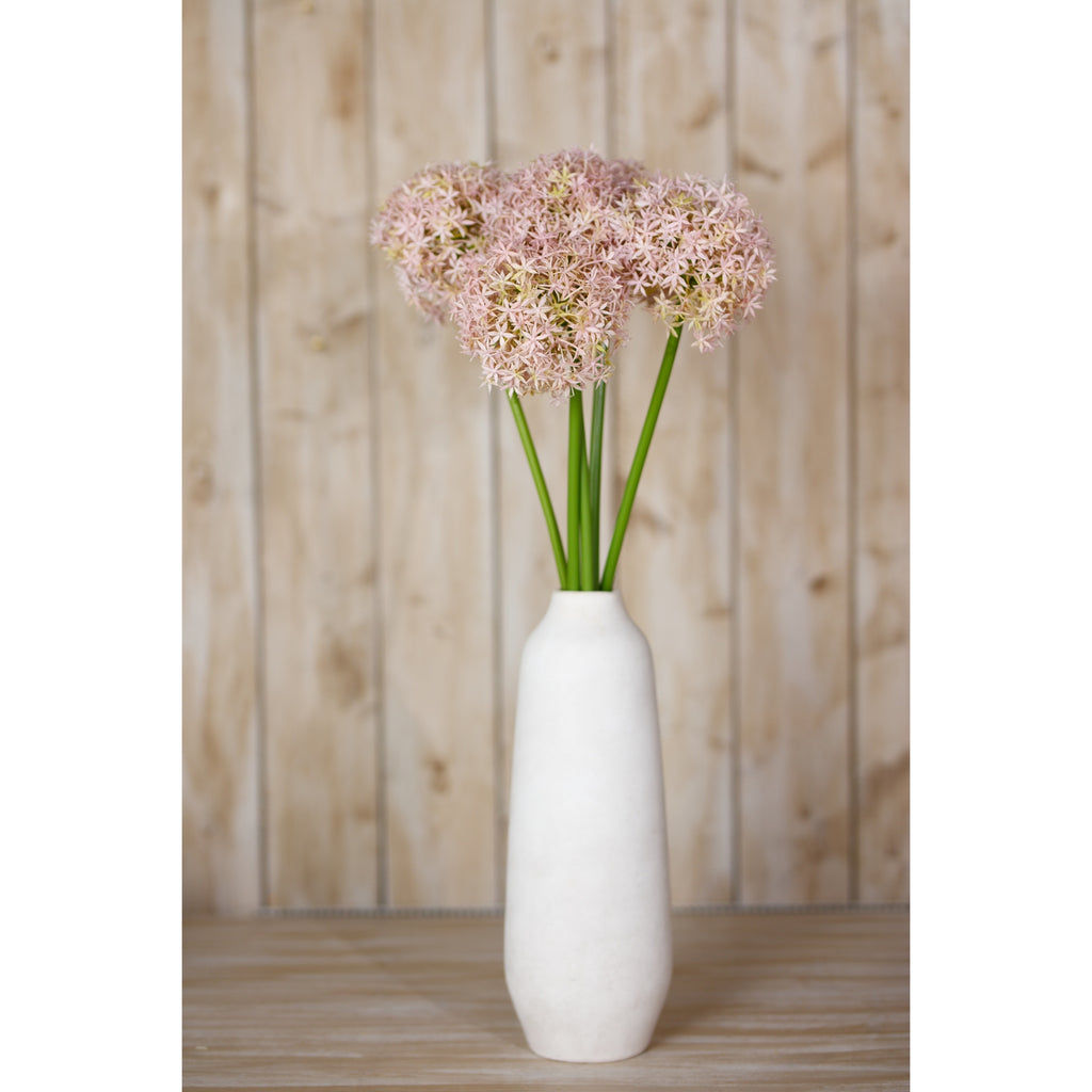 Pale lilac artificial globe flowers arranged in a tall white vase on a wooden surface against timber panelling.
