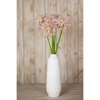 Pale lilac artificial globe flowers arranged in a tall white vase on a wooden surface against timber panelling.