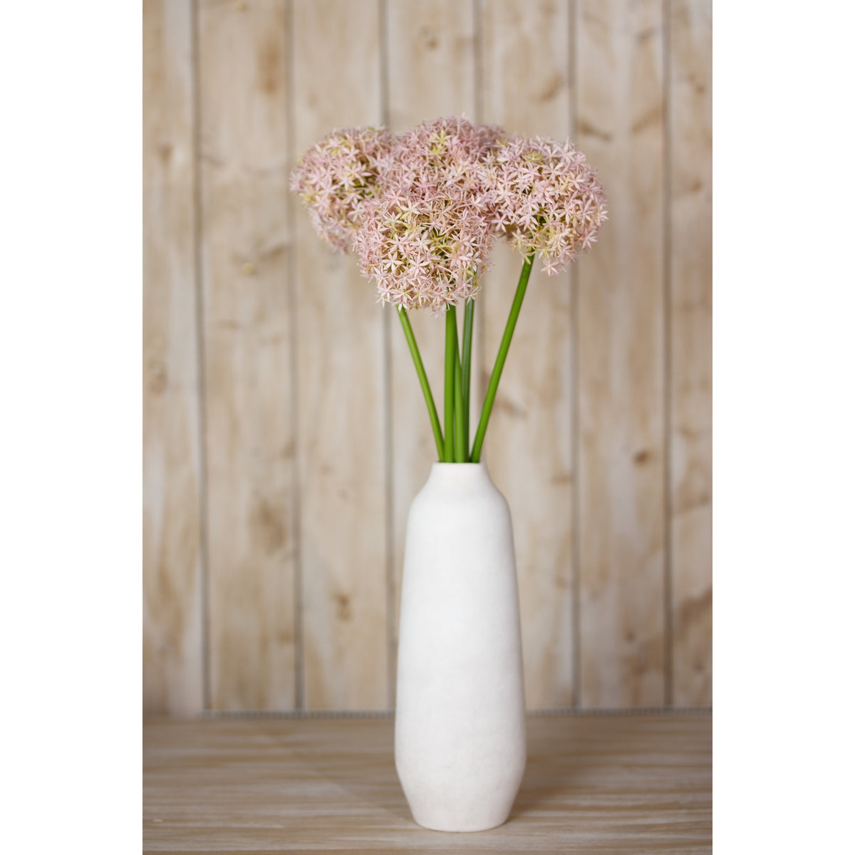 Pale lilac artificial globe flowers arranged in a tall white vase on a wooden surface against timber panelling.