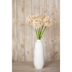 Pale pink artificial globe flowers arranged in a tall white vase on a wooden surface against pale wood panelling.