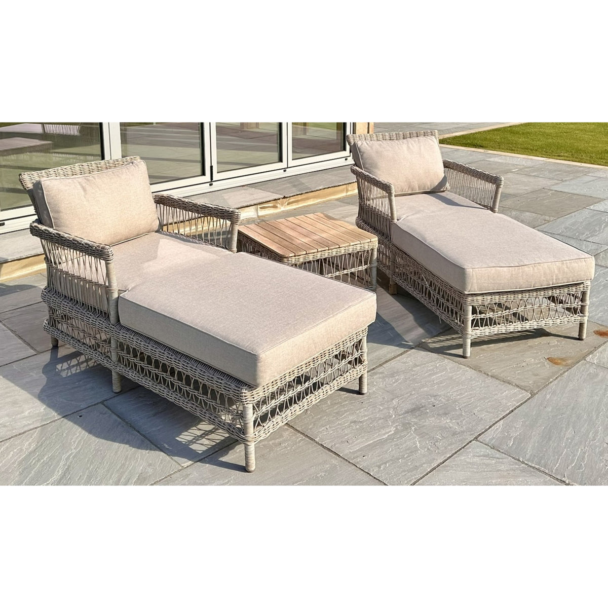 Two pale cushioned woven loungers angled towards the camera on grey paving, with a slatted-top table between them and glass doors behind.