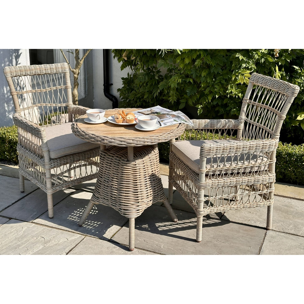 Bistro table with a brown circular top and splayed legs between two beige cushioned chairs, styled with tea and cakes beside leafy planting.