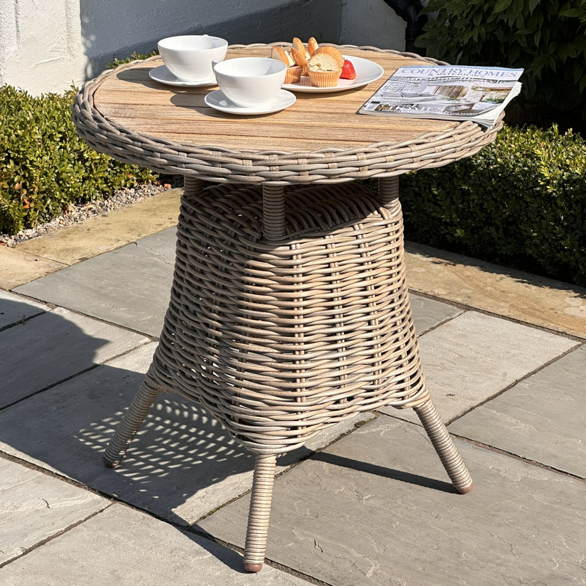 Round bistro table on stone paving, showing an opaque brown top, beige woven-looking rim and tapered base, styled with cups, cakes and a magazine.