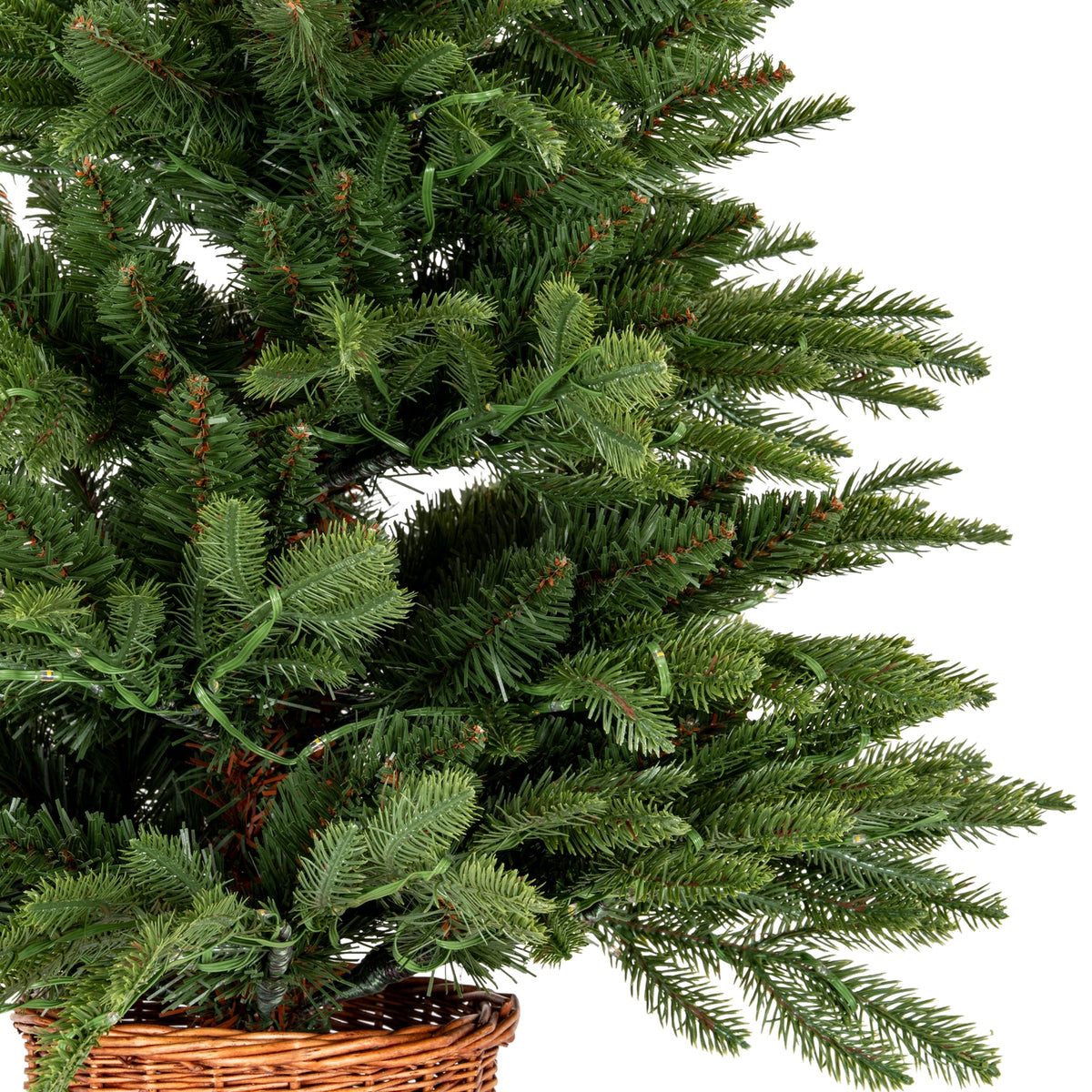 Close-up of dense mixed green artificial fir branches with small clear lamps and green cable threaded through the foliage above the woven pot rim.