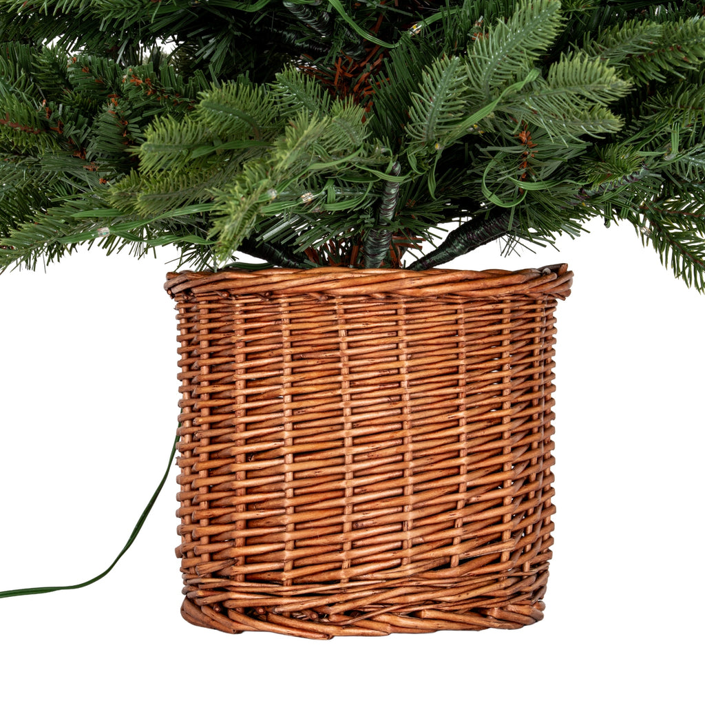 Brown woven pot cover beneath the tree's lower green branches, showing the horizontal weave, wrapped branch stems and trailing green cable.