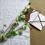 White-flowered artificial branches laid diagonally across pale cloth beside an open notebook with brown ties and a wooden pencil.