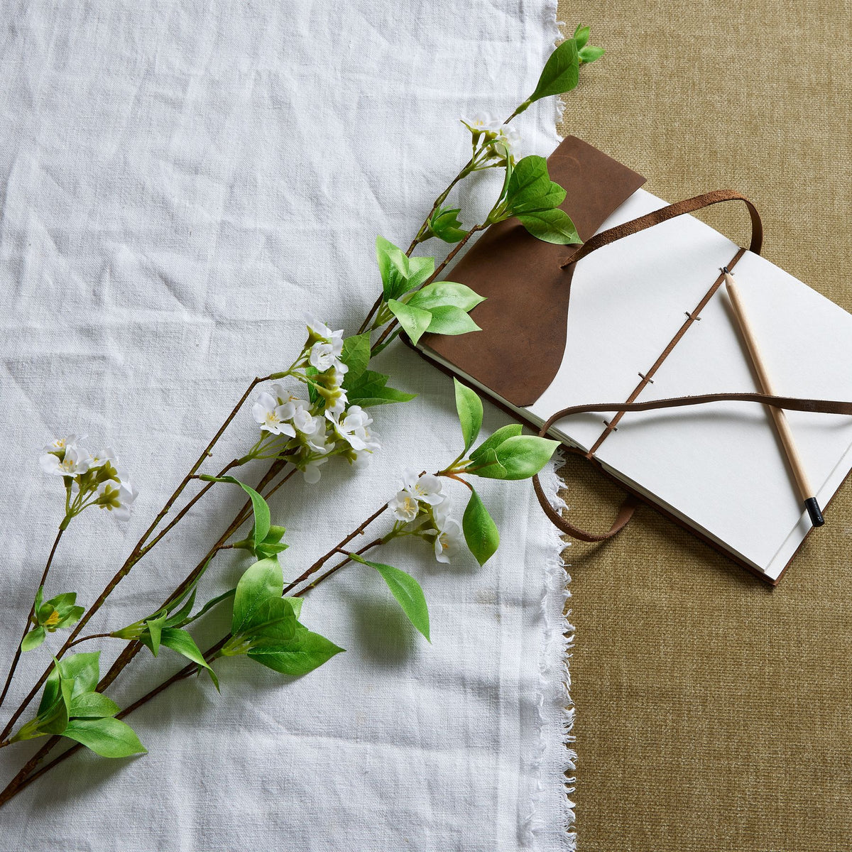 White-flowered artificial branches laid diagonally across pale cloth beside an open notebook with brown ties and a wooden pencil.
